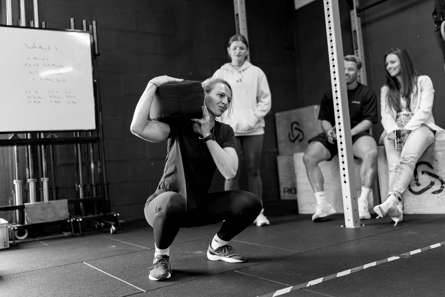 Atleet met sandbag tijdens een squat, crew kijkt toe.